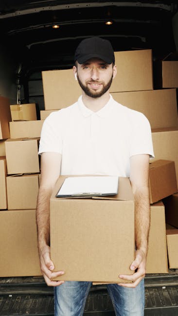 A man wearing a black cap, white t-shirt, and blue jeans is inside a van with an open back, surrounded by neatly stacked cardboard boxes of various sizes. He is holding a medium-sized box in front of