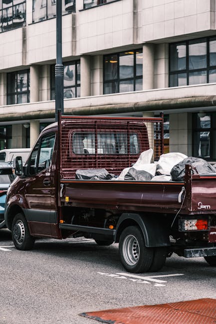 A dark red, small flatbed truck parked on a city street outside a modern office building with large glass windows and white tiled exterior. The truck's bed is loaded with various packed items, includi